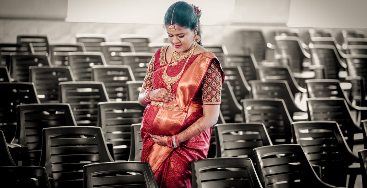 A pregnant woman in a red sari stands in a vast empty auditorium during Carolene's Baby Shower Photoshoot by AK Photography.