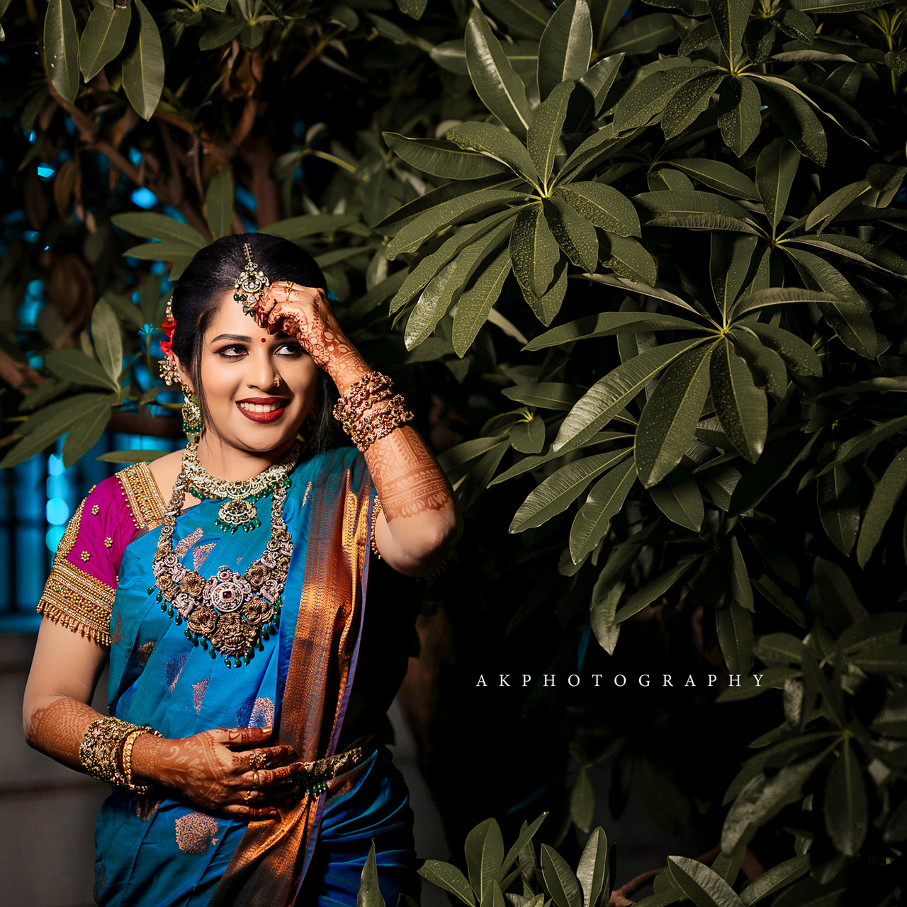 A South Asian woman in her twenties, dressed in a traditional South Indian bridal saree in teal blue and rust gold with intricate embroidery, gold jewelry, and henna designs on her hands. She smiles softly, with a hand gently touching her forehead. Her dark hair is styled in an updo, and she is positioned slightly off-center with a soft-focus green leafy background.