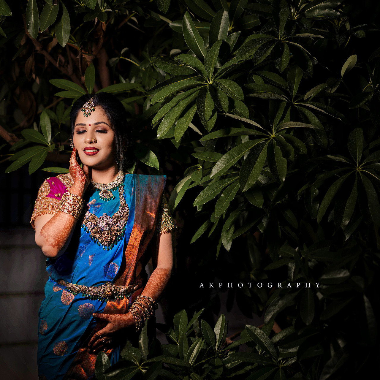 Young South Asian woman in a vibrant sapphire blue South Indian bridal saree with gold and burnt orange accents, gold jewelry, and a serene expression, set against dark green foliage in soft, dramatic lighting.